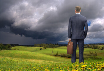 Photoshop illustration. A man in a suit, briefcase in hand, stands in a meadow, watching an approaching storm. Rear view. Stock market crash, crisis, marketing. Business problems. Land trade.  © puchan