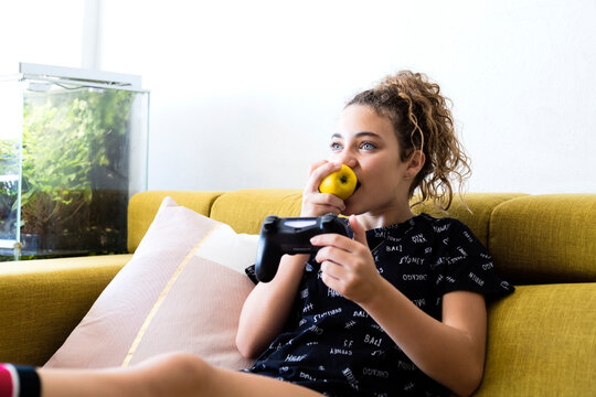 Portrait of girl sitting on the couch at home eating an apple while playing with games consol
