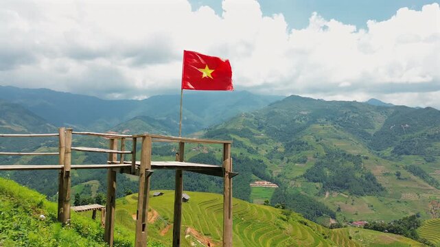 The red flag of Vietnam with its yellow star blows in the wind at a rustic mountain lookout, with lush green terraced fields and misty mountains in the background.