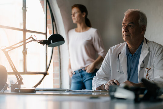 Serious senior doctor sitting at desk in medical practice with woman in background