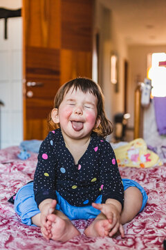 Portrait of toddler sitting barefoot on bed sticking out tongue