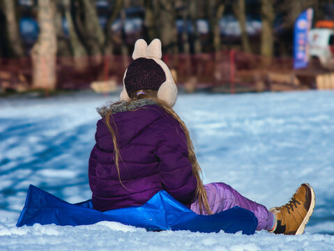 A young girl in a purple jacket and pink pants sits on a blue sledding mat in the snow at Normafa, Budapest. Looking out over the winter park. Perfect for active family and winter tourism themes.