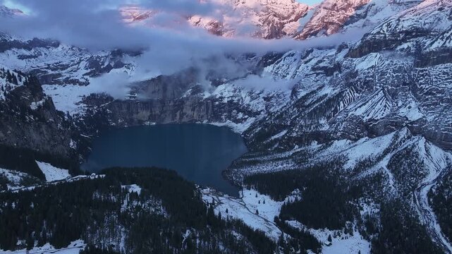 Aerial view of Oeschinensee lake nestled amidst snow-covered mountains under a sky with pink hues, Kandersteg, Canton of Bern, Switzerland.