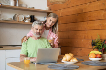 Senior couple using laptop in kitchen while drinking juice