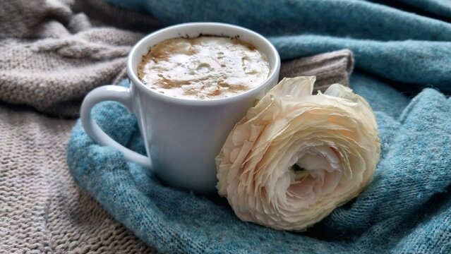 A white cup filled with frothy cappuccino sits on a textured, knitted blue-gray cloth. Beside it lies a large white flower, likely a ranunculus