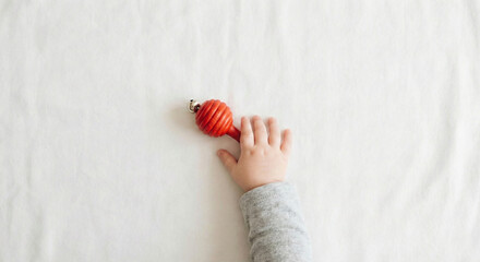Infant hand reaching for red wooden rattle toy on white linen background, baby sensory development concept