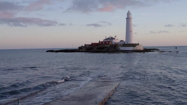 A static medium shot of the historic St Mary&rsquo;s Lighthouse in Whitley Bay at dawn, featuring the white tower and red-roofed cottages on a tidal island with a rotating offshore wind turbine 