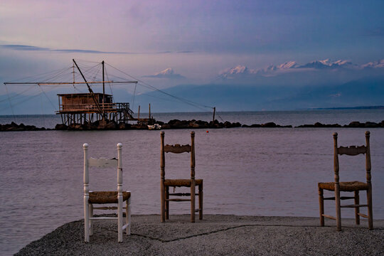 Three wooden chairs facing the sea in the foreground, with a traditional Italian fishing hut (Retone) visible on the water in the distance at Marina di Pisa.