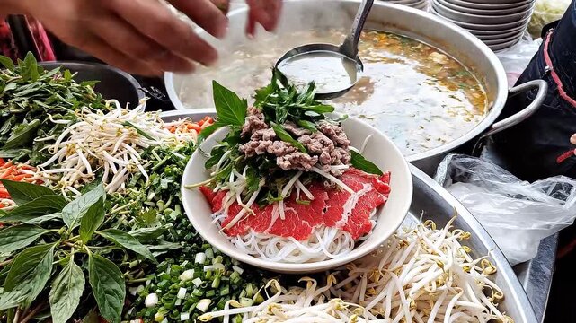 A street vendor is meticulously placing thin slices of raw beef onto a bed of white rice noodles in a bowl, surrounded by fresh basil and bean sprouts