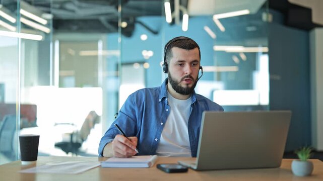 Professional working intently at desk, wearing headset for call. Engaged in online meeting, diligently taking notes. Displays focus, dedication, and concentration in modern office environment.