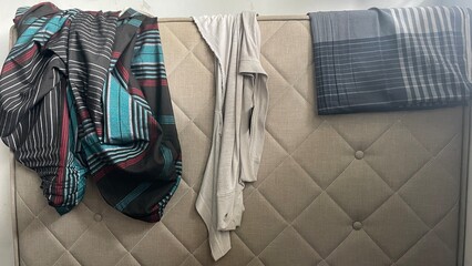 Fototapeta premium Various clothes, including striped and patterned fabrics, and a plain grey garment, casually draped over a padded, tufted headboard, showing textured textiles.