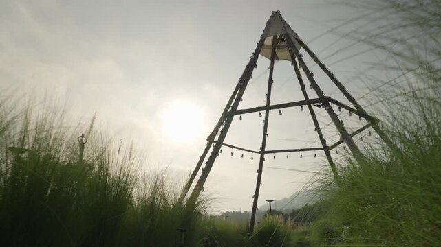 Camera moving forward through lush meadow with sparkling dewdrops towards a wooden tipi and mountain view under a sunstar.