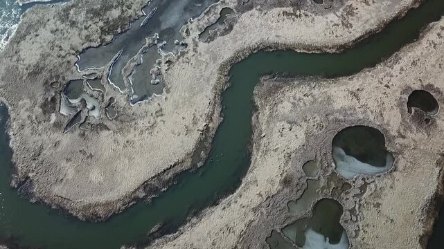 Overhead drone view of a sinuous tidal creek winding through winter salt marsh, with salt pannes, tidal pools and thin ice along the edges, Great Marsh&ndash;Essex Bay, MA