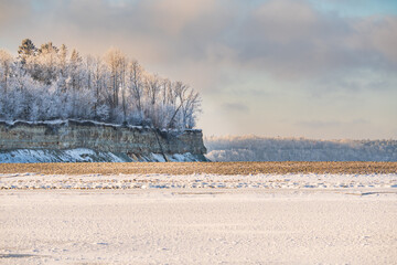 Obraz premium Scenic winter landscape of Estonian limestone cliff at coast with frosted trees and frozen Baltic sea under soft sunset light