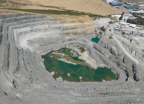Aerial view of a vast quarry carved deep into the earth, revealing layers of grey rock and pools of water, contrasting with the sparse vegetation, Cape Town, Western Cape, South Africa.