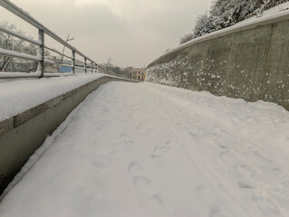 Chodnik jest pokryty śniegiem w zimowy dzień z opadami śniegu. Zaśnieżony chodnik wije się wzdłuż betonowego muru oporowego. Metalowe barierki po lewej stronie wyznaczają granicę ścieżki. © Grzegorz