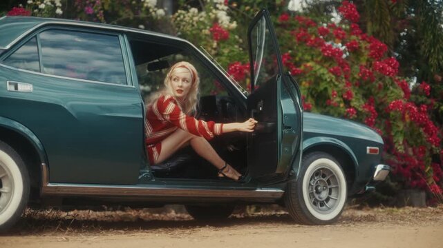 Blonde Woman In Retro Attire By Automobile. Serene Woman Resting Near Vehicle Amidst Lush Flowers