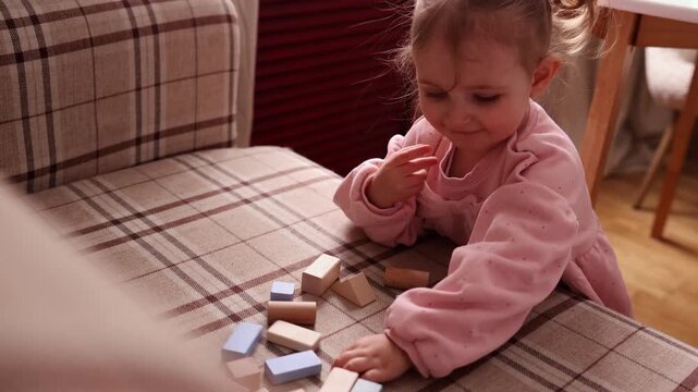 A joyful little girl in a pink dress having fun while breaking a composition of colorful wooden Montessori blocks on a checkered sofa