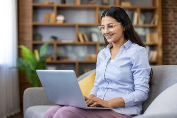 Indian woman wearing eyeglasses and a light blue shirt smiling while typing on a laptop computer, working or studying from a comfortable home couch with bookshelves in the background © Liubomir