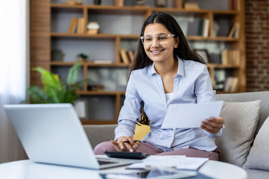 Young indian woman working from her living room, smiling while managing finances using laptop, calculator and paperwork to budget and plan as a remote freelancer entrepreneur