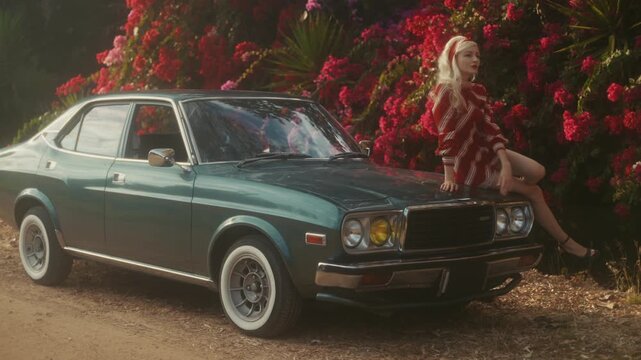Blonde Woman In Retro Attire By Automobile. Serene Woman Resting Near Vehicle Amidst Lush Flowers