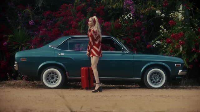 Blonde Woman In Retro Attire By Automobile. Serene Woman Resting Near Vehicle Amidst Lush Flowers