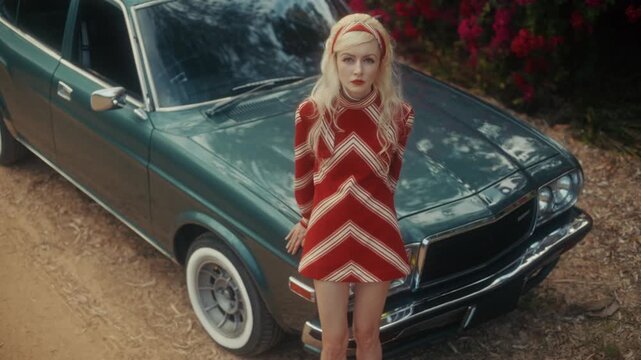 Blonde Woman In Retro Attire By Automobile. Serene Woman Resting Near Vehicle Amidst Lush Flowers
