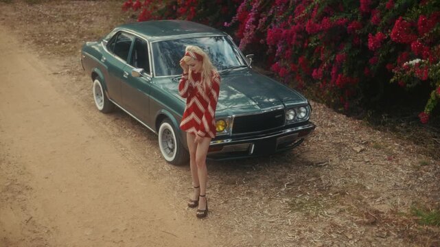 Blonde Woman In Retro Attire By Automobile. Serene Woman Resting Near Vehicle Amidst Lush Flowers