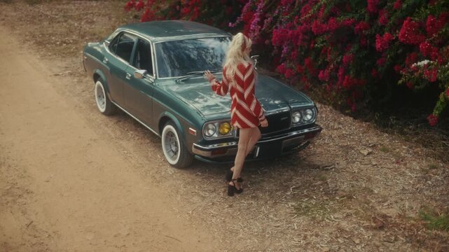Blonde Woman In Retro Attire By Automobile. Serene Woman Resting Near Vehicle Amidst Lush Flowers