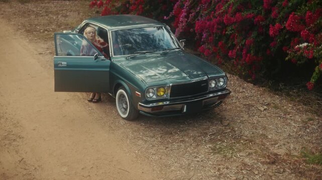 Blonde Woman In Retro Attire By Automobile. Serene Woman Resting Near Vehicle Amidst Lush Flowers