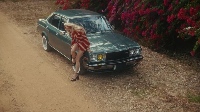 Blonde Woman In Retro Attire By Automobile. Serene Woman Resting Near Vehicle Amidst Lush Flowers