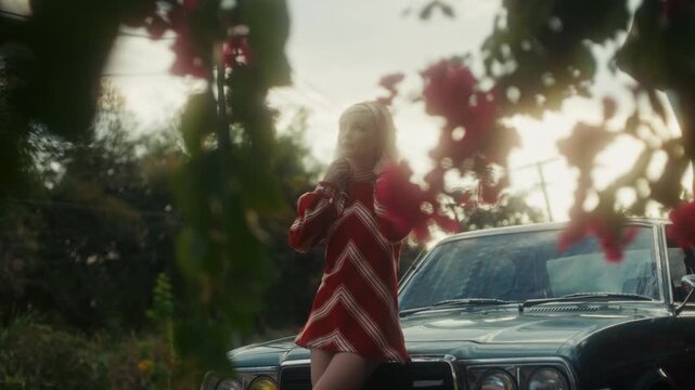 Blonde Woman In Retro Attire By Automobile. Serene Woman Resting Near Vehicle Amidst Lush Flowers