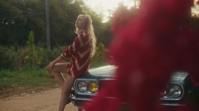 Blonde Woman In Retro Attire By Automobile. Serene Woman Resting Near Vehicle Amidst Lush Flowers
