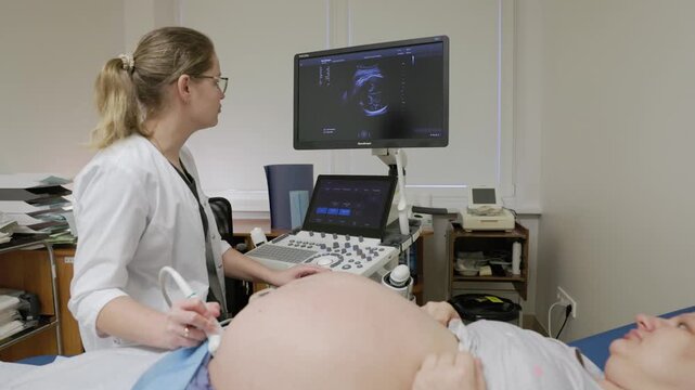 A doctor in a white coat performs an ultrasound scan on a pregnant woman in a clinical setting, with the fetal image visible on the monitor