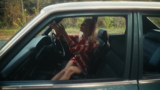 Blonde Woman In Retro Attire By Automobile. Serene Woman Resting Near Vehicle Amidst Lush Flowers