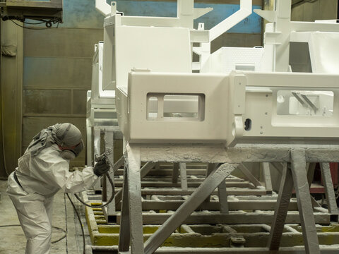 Industrial painter wearing white protective coverall respirator mask gloves spraying liquid coating onto large heavy machinery metal frame parts using professional pneumatic airbrush