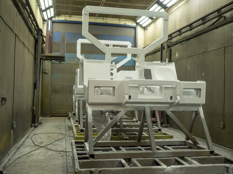 Professional industrial painter wearing white protective coverall suit and respirator mask spraying white liquid coating onto large heavy machinery metal frames using pneumatic airbrush tool