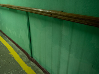 Obraz premium Weathered green textured wall with wooden handrail and yellow safety line painted on concrete floor inside industrial facility or manufacturing plant corridor