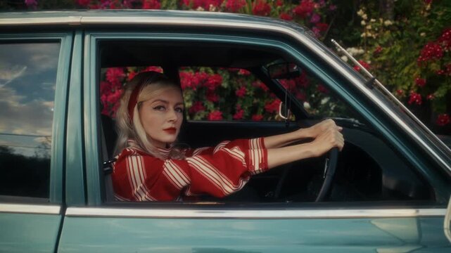 Blonde Woman In Retro Attire By Automobile. Serene Woman Resting Near Vehicle Amidst Lush Flowers