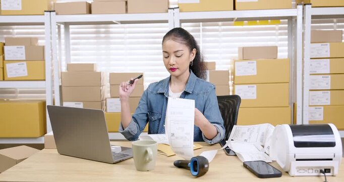 Asian woman entrepreneur checking customer orders from laptop holding shipping document and pen while coordinating online store order fulfillment during small business logistics work inside warehouse