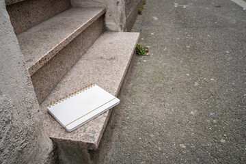 White blank notebook lying on the bottom step outside. Everyday urban scene showing a personal object, representing writing, study, and casual outdoor life.