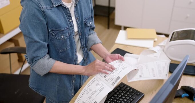 Asian woman small business owner compares printed shipping labels with computer order list, arranges parcel paperwork on desk, manages online store fulfillment and logistics operation