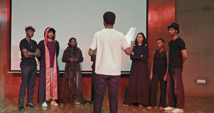 African young adult gen z male man hold scripts paper read play story lines scene standing indoor stage hall. Happy group boy girl do work drama practice day studio campus place