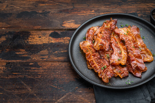 Crispy fried bacon arranged on a black plate over rustic wood offering deep color, shine and high-impact gourmet atmosphere.