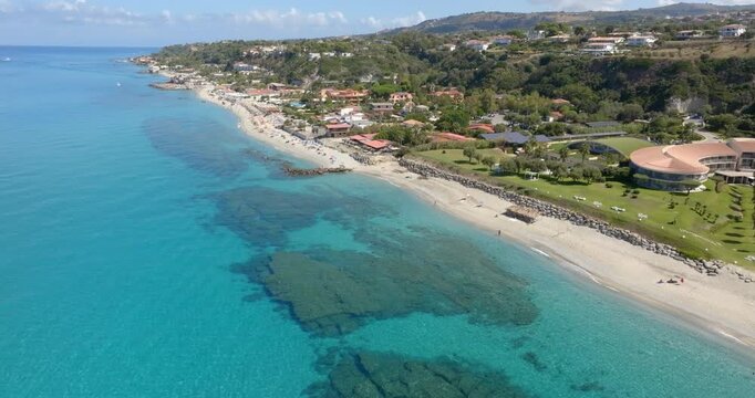 Aerial view of Tono beach, in Italy. A turquoise coastline in Calabria, featuring a white sandy beach, dark reef patches under clear water, and a lush green hillside dotted with Mediterranean villas.