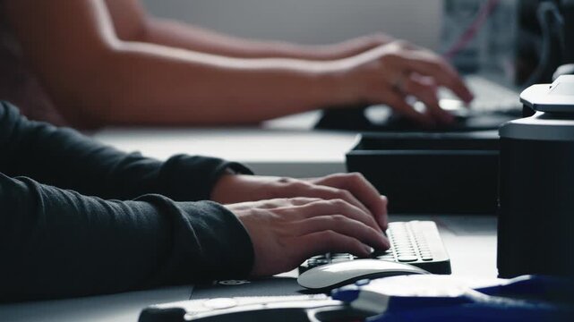 Close-up of male hands typing quickly on a black computer keyboard in a dimly lit modern office. Professional programming or data entry work in a tech startup environment.