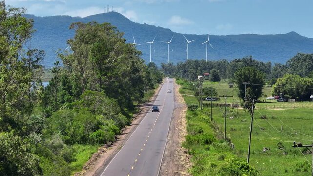 Highway traffic leads through green countryside toward distant wind turbines and mountain ridge, captured in telephoto 7x slow motion with a vertical rise reveal, for ESG energy transition narrative.