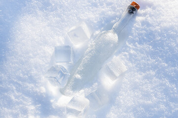 Glass bottle in the fluffy, snowy surface with sunlight surrounded by ice cubes.