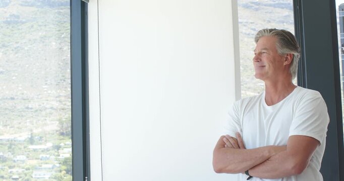Mature man standing on right by window, sunlight drawing gaze, gazing in white tee, copy space