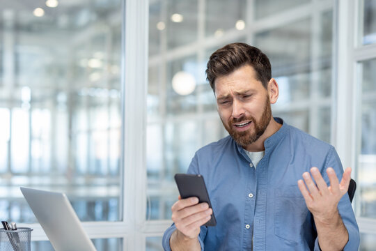 Young man feeling confused and frustrated, holding a smartphone in an office setting, trying to solve a technical problem or dealing with a bad connection